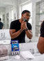 A man in a black t-shirt shaving in a marble bathroom, with a boxed 4-blade razor kit on the counter in front of him.