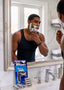 A man in a black tank top shaves in a bright bathroom, with a boxed 6-blade razor and several loose razor cartridges placed on the sink counter in the foreground.