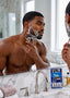 A shirtless man shaving in a bathroom mirror while holding a boxed shaving kit on the counter. The shaving kit contains multiple razors and is displayed next to a chrome faucet and marble countertop.