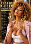 A woman with voluminous curly hair holds two gold-finished afro hair picks branded SPI STYLES. She is wearing a pink and white checkered blazer, a white crop top, and gold jewelry against a warm, blurred background.