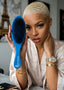 A woman with a buzz cut holds up a blue hairbrush with dark bristles. The brand name 'SPI STYLES' is printed on the handle.