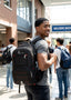 A man wearing a black and red Victor backpack stands in a school hallway, smiling and looking to the side. Other students and a brick building are visible in the background.