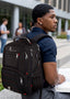 A man wearing a navy blue polo shirt and gray pants sits outdoors on a stone ledge, carrying a large black laptop backpack with multiple zippered compartments and red accent stitching. The backpack has a white 'GOT FLYING' logo on the upper left side.