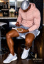 A man wearing a light pink hoodie, gray shorts, and white sneakers sits on a white bar stool in a modern kitchen setting, holding a white paddle brush with a gold bristle head. The SPI STYLES logo is visible in the bottom right corner.