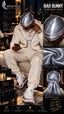 A man wearing a silver holographic 'Bad Bunny' silk durag and a matching cream-colored tracksuit sits on a leather couch in a modern living room setting. The right side of the image features a multi-panel infographic detailing the product's features, including close-ups of the silver fabric texture and the back view of the durag.