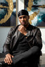 Man in a leather jacket and black cap sitting in a chair with abstract art in the background