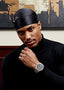 Man wearing a black turtleneck and black headwrap with a watch, standing against a textured wall.