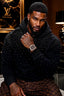 A man wearing a black Louis Vuitton hoodie covered in sparkling rhinestones and matching brown monogram pants, posed in a luxury interior with Louis Vuitton branding visible in the background.