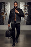 Man in a stylish outfit with a black jacket and pants, holding a black bag in an art gallery setting.