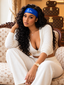 A woman with long, curly black hair sits on a patterned sofa wearing a white ribbed knit two-piece outfit and a bright blue headband with a white logo. The background features vintage Louis Vuitton-style monogram trunks.