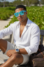Man wearing a white shirt and sunglasses sitting outdoors with greenery in the background