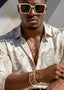 Man wearing a patterned shirt, sunglasses, and gold jewelry against a neutral background