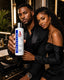 A man and a woman pose together in a luxurious bathroom setting, with the man holding up a bottle of Spistyles Haircare product. The man is wearing a black suit, and the woman is wearing a black off-the-shoulder dress.