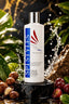 A bottle of SPISTYLES Clearwater Coconut Botanical Oil stands on a black marble pedestal, surrounded by tropical leaves, macadamia nuts, and red berries with water splashes in the background.