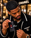 A man in a black Chanel jacket holds up a black wallet featuring the 'SPI STYLES' logo. The background shows a blurred Chanel store interior.
