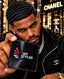 A man in a black Chanel jacket holds up a black leather wallet featuring the 'SPI STYLES' logo. The background shows a blurred Chanel store interior with gold lights and logos.