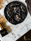 A black satin hair accessory with a gold geometric animal face pattern, styled on a white marble surface next to a bottle of hair essence and a branded box.