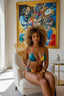 Woman in a colorful bikini sitting on a chair holding a comb, with a vibrant painting in the background.