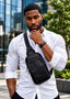A man wearing a black textured leather crossbody sling bag over a white button-down shirt, standing outdoors in front of a modern glass building.