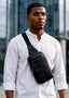 A man wearing a white long-sleeve shirt and a black leather crossbody sling bag with a black strap, standing in front of a modern glass building.