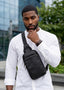A man wearing a white long-sleeve shirt and a black leather crossbody sling bag, standing outdoors in front of a modern glass building.