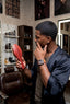 Man in a barber shop holding a red hairbrush, with shelves and mirrors in the background.