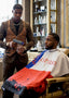 Barber in a suit with apron attending to a customer in a barber chair, with shelves of products in the background.
