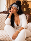 A woman with long, curly black hair sits on a Louis Vuitton-patterned sofa, wearing a white ribbed knit two-piece set with a deep V-neck and front zipper, a gray headband with 'STYLES' printed on it, and a gold watch.