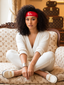 A woman with curly black hair sits cross-legged on a beige Louis Vuitton monogram sofa, wearing a red headband with a white LV logo, a cream ribbed cardigan, cream pants, and white loafers with gold hardware. Louis Vuitton luggage is visible in the background.