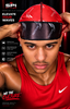 A man wearing a red and black patterned SPI Styles premium silk curag (wavy hair cap) and a red Nike tank top, posing with his hands on his head against a dark red background with promotional text.