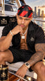 A man sitting on a yacht wearing a black and red striped SPI Styles bandana, a black short-sleeve button-up shirt with SPI Styles embroidery, and matching black shorts. He is accessorized with a gold chain necklace, a silver bracelet, and a gold watch, with a gold SPI Styles cup in the foreground.