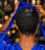 A man viewed from behind wearing a blue satin cap with a black leather patch that reads 'SPI STYLES' in white, with his hands lifting the cap to reveal his styled black hair. He is wearing a blue shirt and a silver chain necklace.