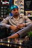 A man wearing a shiny silver-gray silk durag and matching trousers, sitting in a leather chair in a luxury boutique. He is wearing a vibrant blue and green patterned silk headwrap and multiple gold chains.